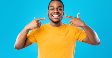 Look At My Smile. Happy Black Man Pointing Fingers At His Mouth Smiling Showing Perfect White Teeth Over Blue Studio Background, Wearing Yellow T-Shirt. Stomatology And Toothcare Concept