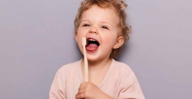 Portrait of laughing happy cheerful infant girl kid with curly blonde hair brushing her teeth, taking care of dental hygiene standing isolated over gray background.