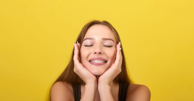 The image shows a young woman with long brown hair and braces on her teeth. She is wearing a black tank top and has her hands on her face. The background is a bright yellow color.